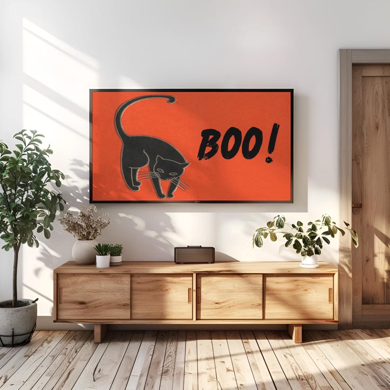 May include: A black cat illustration on an orange background with the word "BOO!" in black. The artwork is displayed on a wall-mounted television above a wooden cabinet. The room has a wooden floor and decorative plants.