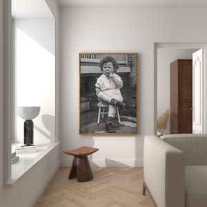 Peut inclure: Photographie en noir et blanc d'un jeune enfant assis sur un tabouret en bois. L'enfant porte une chemise &agrave; manches courtes et un short. L'enfant regarde la cam&eacute;ra et a le doigt dans la bouche.