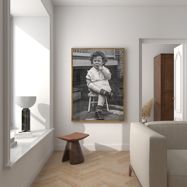 Peut inclure: Photographie en noir et blanc d'un jeune enfant assis sur un tabouret en bois. L'enfant porte une chemise &agrave; manches courtes et un short. L'enfant regarde la cam&eacute;ra et a le doigt dans la bouche.