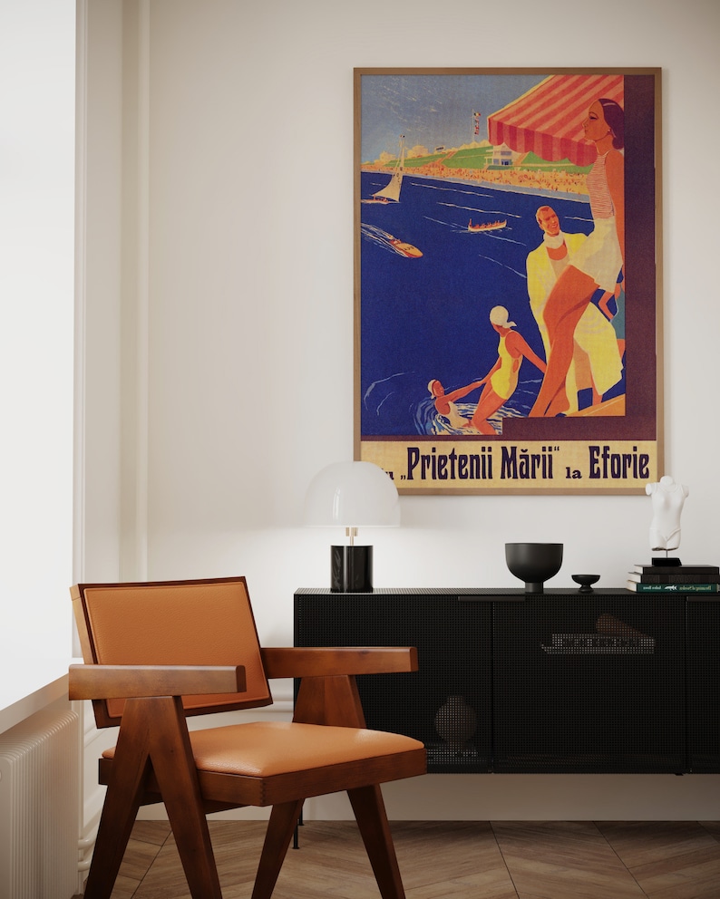 May include: A mid-century modern chair with a brown wooden frame and a tan leather seat. A vintage travel poster in a wooden frame hangs on the wall. The poster features a beach scene with people and the text "Prietenii Mării la Eforie".