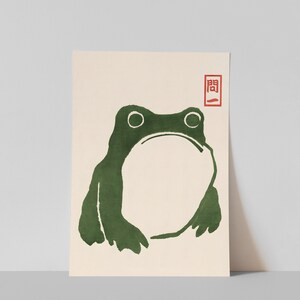May include: A green frog illustration with a simple, minimalist style. The frog is facing forward with its mouth closed and its eyes wide open. The illustration is on a cream-colored background with a red stamp that says "関".