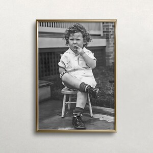 Peut inclure: Une photo en noir et blanc d'un jeune enfant assis sur une petite chaise en bois. L'enfant porte une chemise blanche et un short et tient un petit objet dans sa bouche. La photo est encadr&eacute;e dans un cadre dor&eacute;.