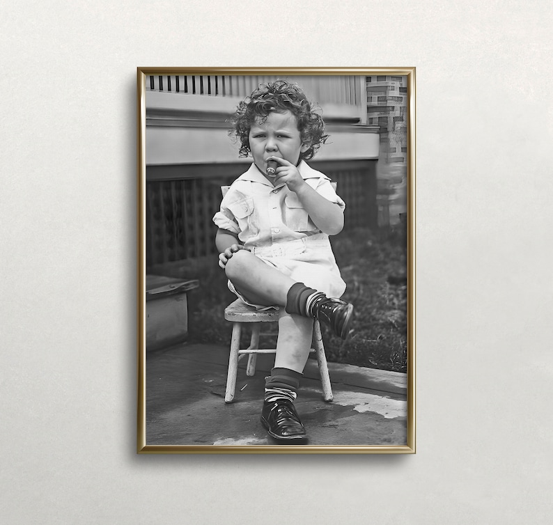 Peut inclure: Une photo en noir et blanc d'un jeune enfant assis sur une petite chaise en bois. L'enfant porte une chemise blanche et un short et tient un petit objet dans sa bouche. La photo est encadr&eacute;e dans un cadre dor&eacute;.