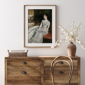 May include: A framed print of a classic portrait. The artwork shows a woman in a long, flowing white dress, seated on a red chair. The print is displayed in a wooden frame, adding a touch of elegance to the room's decor.