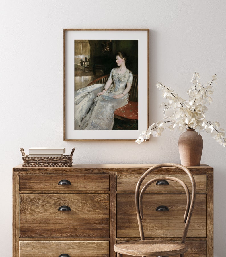 May include: A framed print of a classic portrait. The artwork shows a woman in a long, flowing white dress, seated on a red chair. The print is displayed in a wooden frame, adding a touch of elegance to the room's decor.