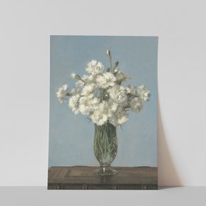 以下が含まれることがあります： 透明なガラスの花瓶に入った白い花の絵。花は密集しており、いくつかのつぼみが見えます。花瓶は、明るい青色の背景に対して、暗い木製の表面に置かれています。