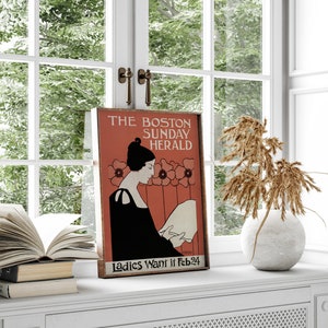 May include: A framed vintage poster for The Boston Sunday Herald, showing a woman in black reading a paper, set against a red background with floral accents. The text "Ladies Want it Feb 24" is visible.