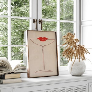 May include: A framed rectangular wall art print featuring a minimalist red line drawing of a martini glass with a pair of bright red lips positioned at the rim. The artwork is set against a light beige background and enclosed in a wooden frame.