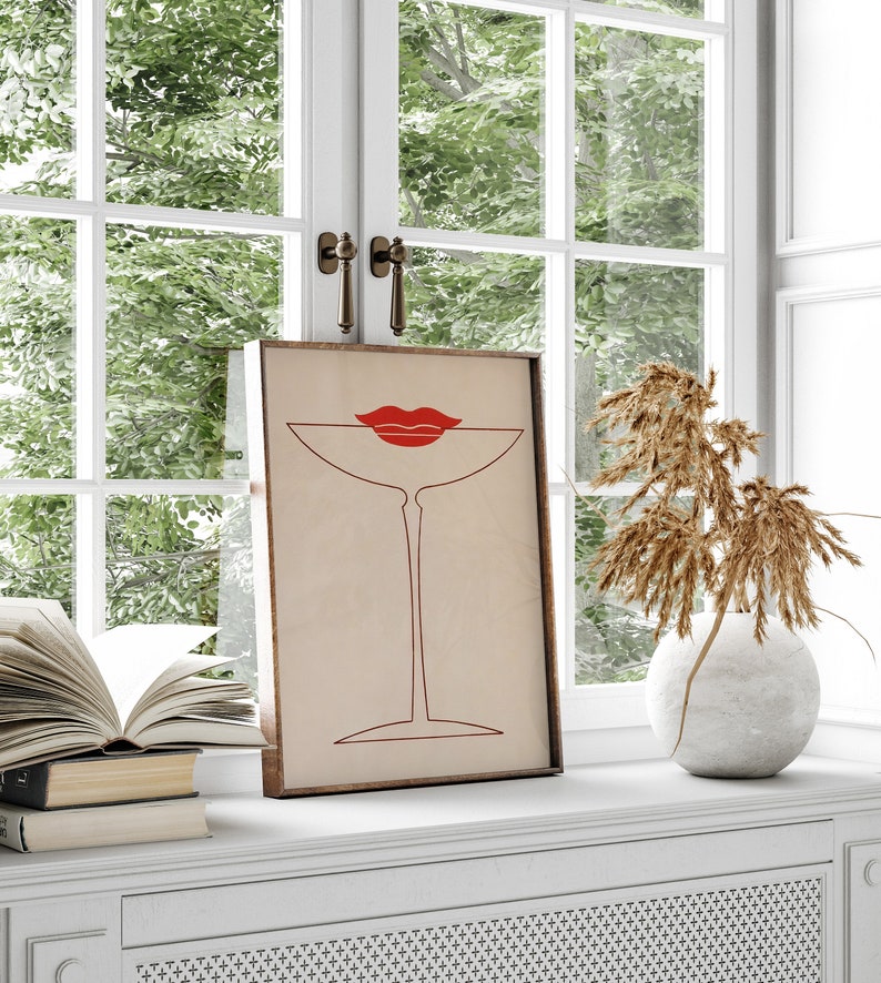 May include: A framed rectangular wall art print featuring a minimalist red line drawing of a martini glass with a pair of bright red lips positioned at the rim. The artwork is set against a light beige background and enclosed in a wooden frame.