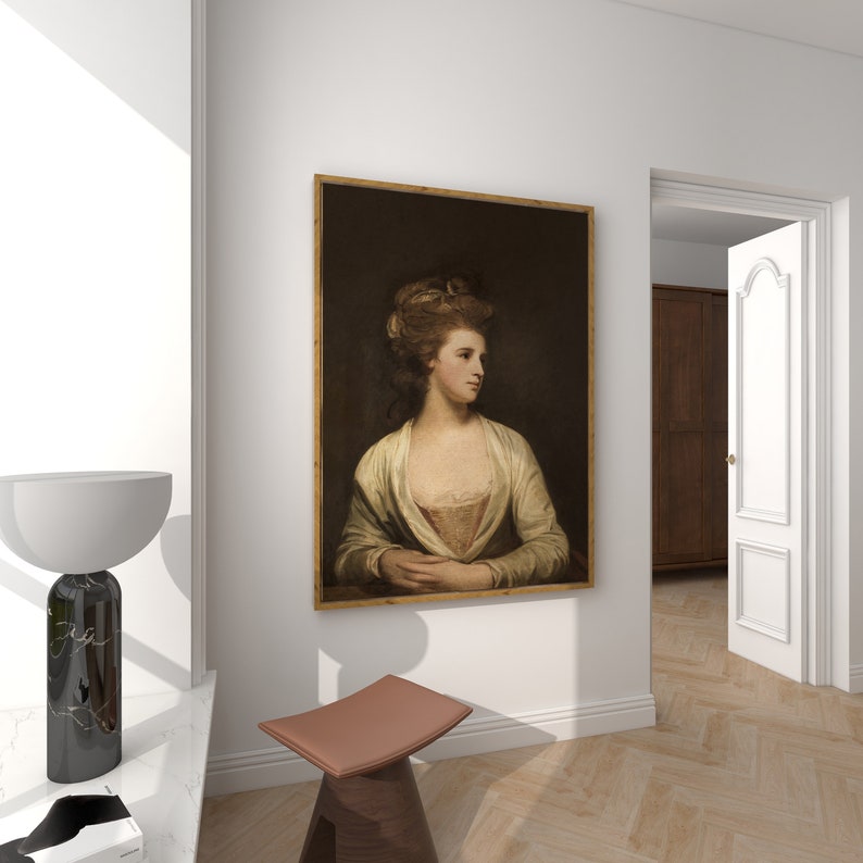May include: A portrait of a woman in a cream dress is displayed on a white wall, framed in a gold-toned border. The artwork shows a woman with fair skin and her hands clasped. A modern lamp and stool are in the foreground.