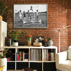 May include: Black and white photograph of five women playing golf on a grassy field. The women are wearing 1920s clothing and are standing on blocks of ice. The photograph is framed in a gold frame.