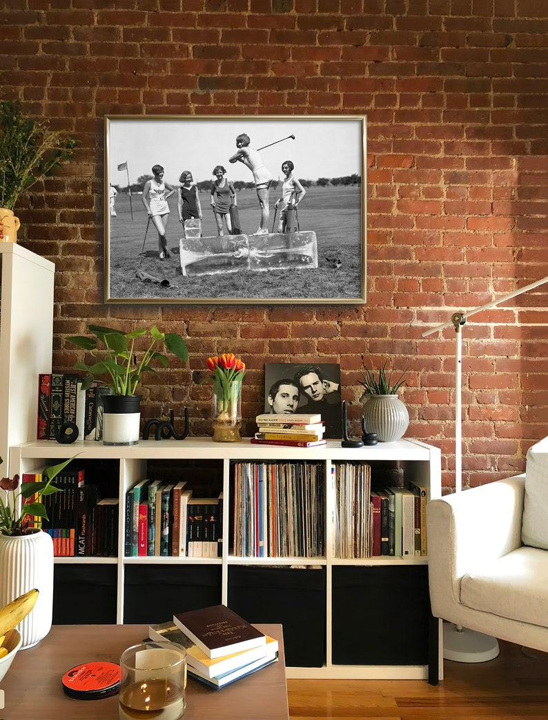 May include: Black and white photograph of five women playing golf on a grassy field. The women are wearing 1920s clothing and are standing on blocks of ice. The photograph is framed in a gold frame.
