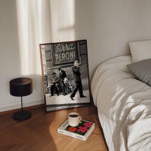 Pu&ograve; includere: Una fotografia in bianco e nero incorniciata con la scritta "birra PERONI" sopra tre uomini in abito. Una tazza di caff&egrave; &egrave; appoggiata su un libro con fiori rossi e rosa. Sono visibili anche una lampada nera e un letto con lenzuola bianche.