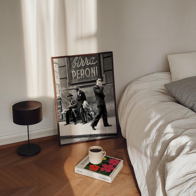 Pu&ograve; includere: Una fotografia in bianco e nero incorniciata con la scritta "birra PERONI" sopra tre uomini in abito. Una tazza di caff&egrave; &egrave; appoggiata su un libro con fiori rossi e rosa. Sono visibili anche una lampada nera e un letto con lenzuola bianche.