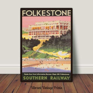 May include: A vintage travel poster for Folkestone, showing a large white building with orange awnings under a pink sky. The poster reads "FOLKESTONE" at the top and "SOUTHERN RAILWAY" at the bottom, with a beach scene in the foreground. The poster is framed.
