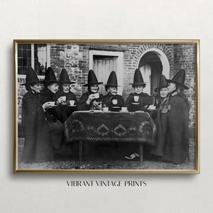 May include: A vintage black and white photograph of seven women dressed in black witch costumes and hats, sitting around a table with teacups. They are all smiling and looking at the camera. The photo is set in front of a brick building.