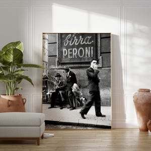 Pu&ograve; includere: Stampa incorniciata in bianco e nero raffigurante una scena di strada con uomini e un'insegna con la scritta "Birra Peroni". La stampa ha una cornice dorata ed &egrave; esposta in una stanza con una pianta, un pouf e vasi di terracotta.