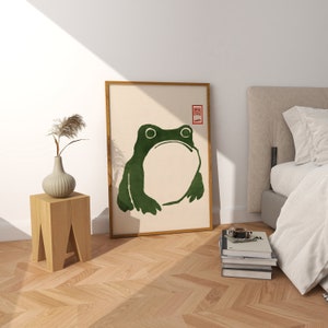 May include: A framed print of a green frog with a white background. The frog is facing forward with its mouth closed. The print has a red stamp in the top right corner with the Japanese character "関" (kan).