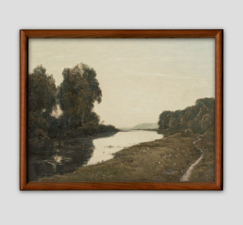 May include: A framed landscape painting depicting a river winding through a forested area. The artwork features muted tones of green, brown, and gray, with trees lining the riverbanks and a path along the shore. The frame is a dark wood.