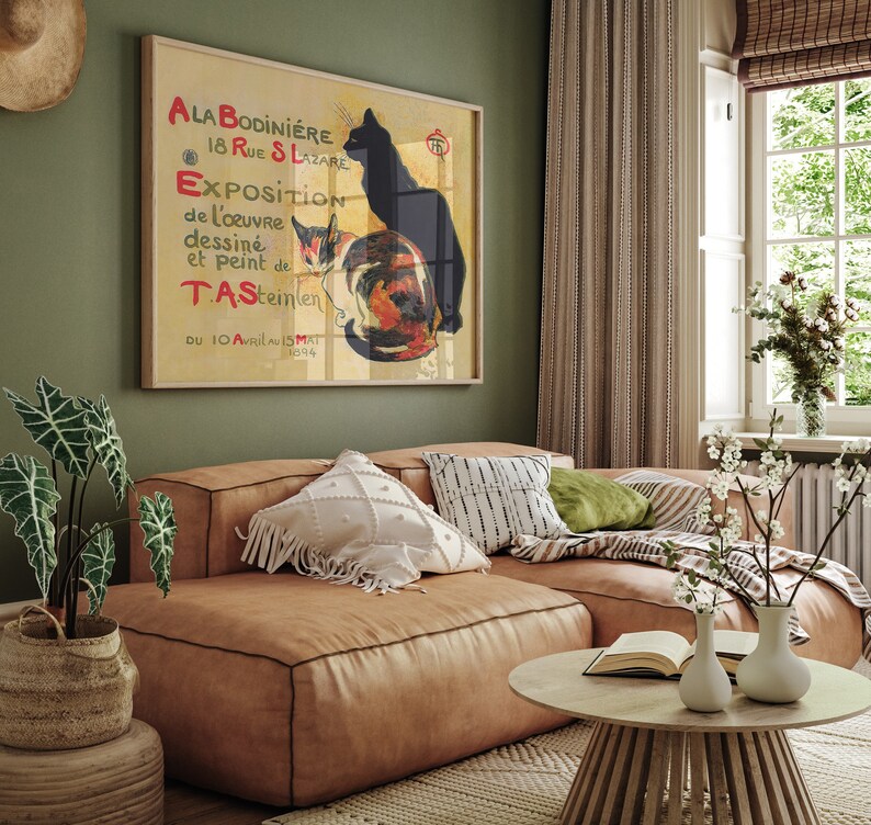 May include: A framed vintage poster featuring a black cat and a calico cat, with French text. The poster is displayed above a brown leather sectional sofa. The room has a neutral color palette with green walls and a wooden coffee table.