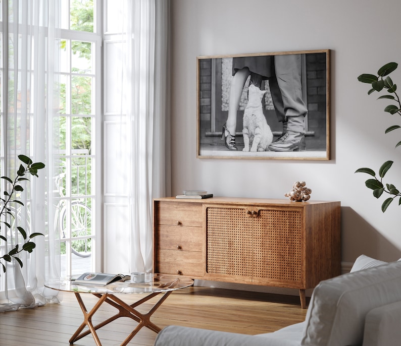 K&ouml;nnte beinhalten: Ein Holzschr&auml;nkchen mit einer geflochtenen Rattan-T&uuml;r und drei Schubladen. Der Schrank steht in einem Wohnzimmer mit einem wei&szlig;en Sofa, einem Glas-Couchtisch und einem Schwarzwei&szlig;foto eines Paares und einer Katze, das an der Wand h&auml;ngt.