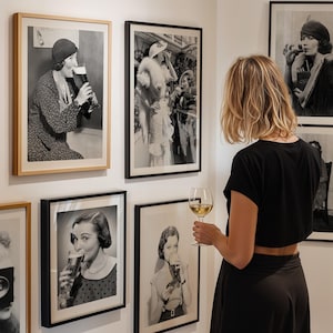 May include: A woman wearing a black dress and a white shirt is looking at a framed black and white photograph of a woman in a fur coat and hat. The photograph is hanging on a white wall. There are other framed black and white photographs on the wall.