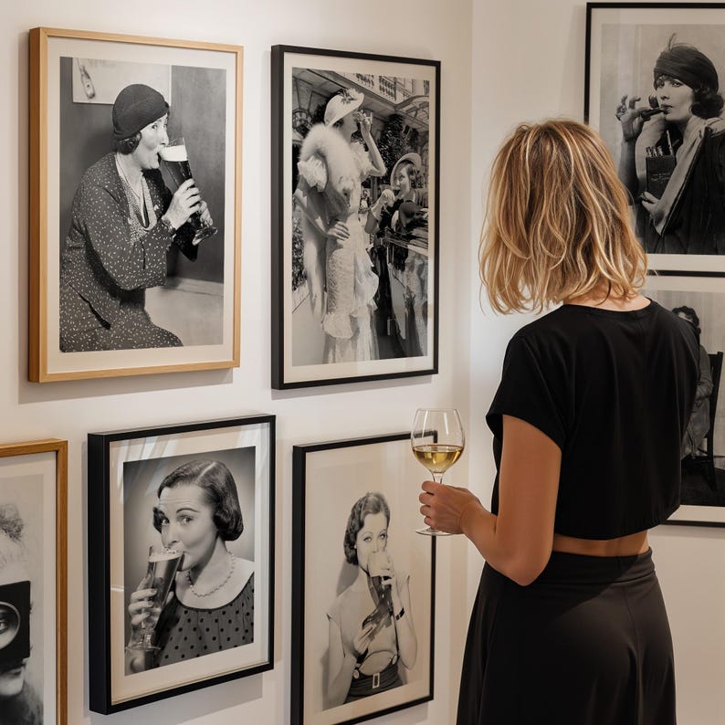 May include: A woman wearing a black dress and a white shirt is looking at a framed black and white photograph of a woman in a fur coat and hat. The photograph is hanging on a white wall. There are other framed black and white photographs on the wall.