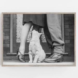 K&ouml;nnte beinhalten: Ein Schwarzwei&szlig;foto von einem Paar, das mit einer Katze dazwischen steht. Das Paar tr&auml;gt Kleidung aus den 1950er Jahren. Die Katze schaut das Paar an.