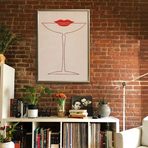 May include: A framed wall art print featuring a minimalist red line drawing of a martini glass with a pair of red lips resting on the rim. The artwork is set against a light cream background and enclosed in a thin, metallic silver frame.