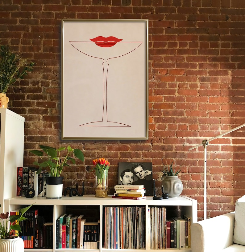 May include: A framed wall art print featuring a minimalist red line drawing of a martini glass with a pair of red lips resting on the rim. The artwork is set against a light cream background and enclosed in a thin, metallic silver frame.