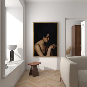 Pode incluir: Um retrato emoldurado de uma mulher com cabelo escuro e um vestido verde, sobre um fundo escuro. A pintura est&aacute; pendurada em uma parede branca em uma sala com piso de madeira e um sof&aacute; branco.