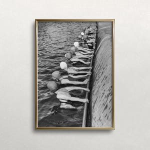 May include: Black and white framed photograph of swimmers lined up at the edge of a pool or lake. The swimmers are wearing swim caps and swimsuits, ready to dive into the water. The image has a vintage, documentary feel.