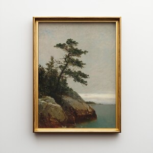 May include: A framed landscape painting depicting a lone pine tree growing on a rocky shoreline overlooking a calm body of water under a cloudy, overcast sky. The artwork is enclosed in a thin, metallic gold frame with a simple, clean profile.