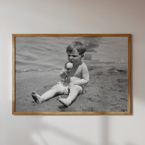 May include: A black and white photo of a young boy sitting on a sandy beach, eating an ice cream cone.