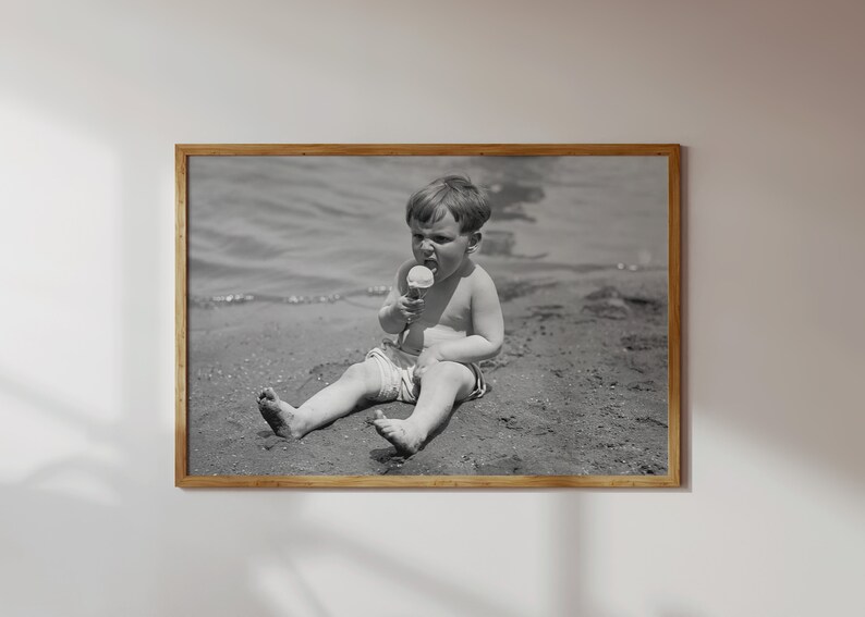 May include: A black and white photo of a young boy sitting on a sandy beach, eating an ice cream cone.