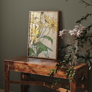 May include: A framed botanical print of yellow flowers with green leaves on a beige background. The print is hanging on a dark green wall above a wooden table.