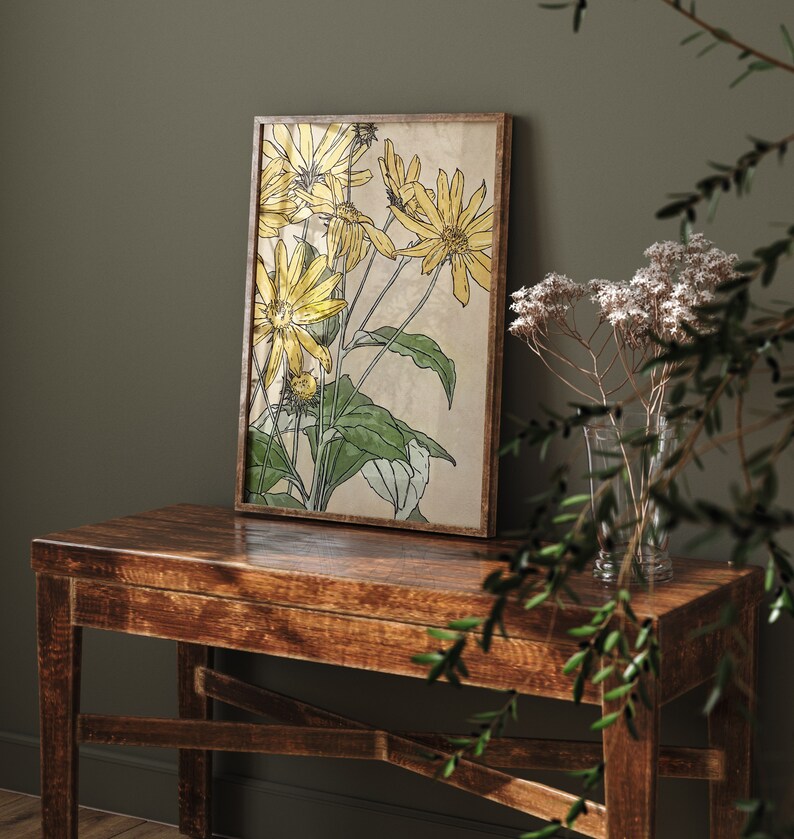 May include: A framed botanical print of yellow flowers with green leaves on a beige background. The print is hanging on a dark green wall above a wooden table.
