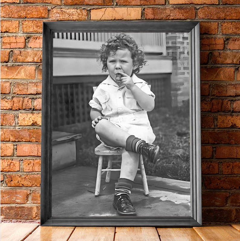 Peut inclure: Une photo en noir et blanc d'un jeune enfant assis sur une chaise en bois. L'enfant porte une chemise blanche et un short et a les cheveux boucl&eacute;s. L'enfant tient un petit objet dans sa bouche.