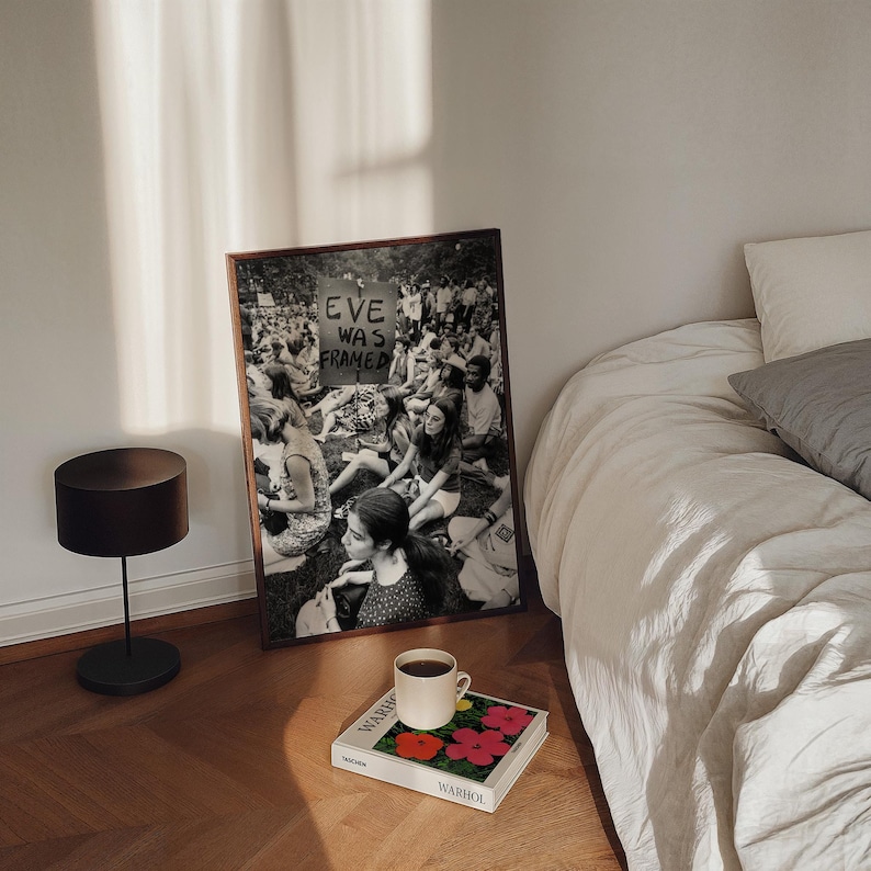 Puede incluir: Una fotograf&iacute;a en blanco y negro enmarcada de una multitud de personas que sostienen un cartel que dice "Eve Was Framed". La fotograf&iacute;a est&aacute; colgada en una pared blanca junto a una cama con s&aacute;banas blancas y una almohada gris. Una l&aacute;mpara de mesa negra est&aacute; en el suelo junto a la fotograf&iacute;a. Un libro con un dise&ntilde;o de flores coloridas y la palabra "Warhol" en la portada est&aacute; sobre la cama.