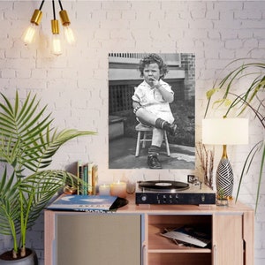 Peut inclure: Une photographie en noir et blanc d'un jeune enfant assis sur un petit tabouret en bois. L'enfant porte une robe blanche et a les cheveux boucl&eacute;s. L'enfant regarde la cam&eacute;ra et a une expression s&eacute;rieuse sur son visage.