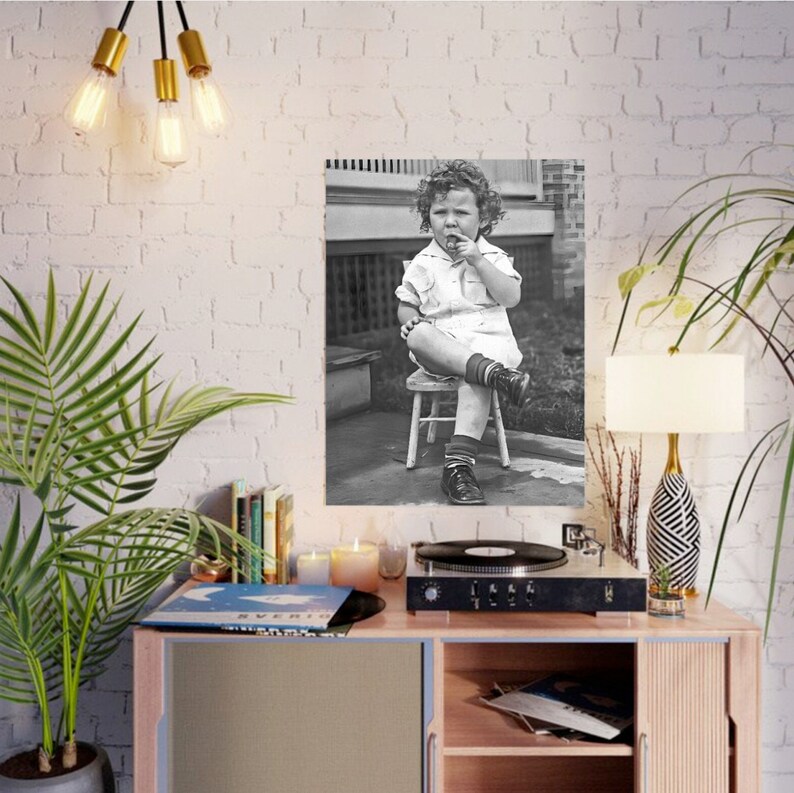 Peut inclure: Une photographie en noir et blanc d'un jeune enfant assis sur un petit tabouret en bois. L'enfant porte une robe blanche et a les cheveux boucl&eacute;s. L'enfant regarde la cam&eacute;ra et a une expression s&eacute;rieuse sur son visage.