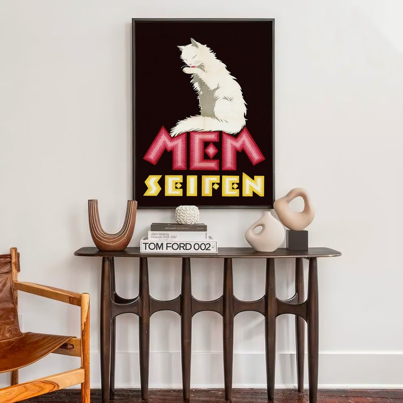 May include: A framed art print featuring a white cat grooming itself against a dark background. The print has the text "MEM SEIFEN" in pink and yellow. The print is displayed above a dark wood console table with decorative vases and books.
