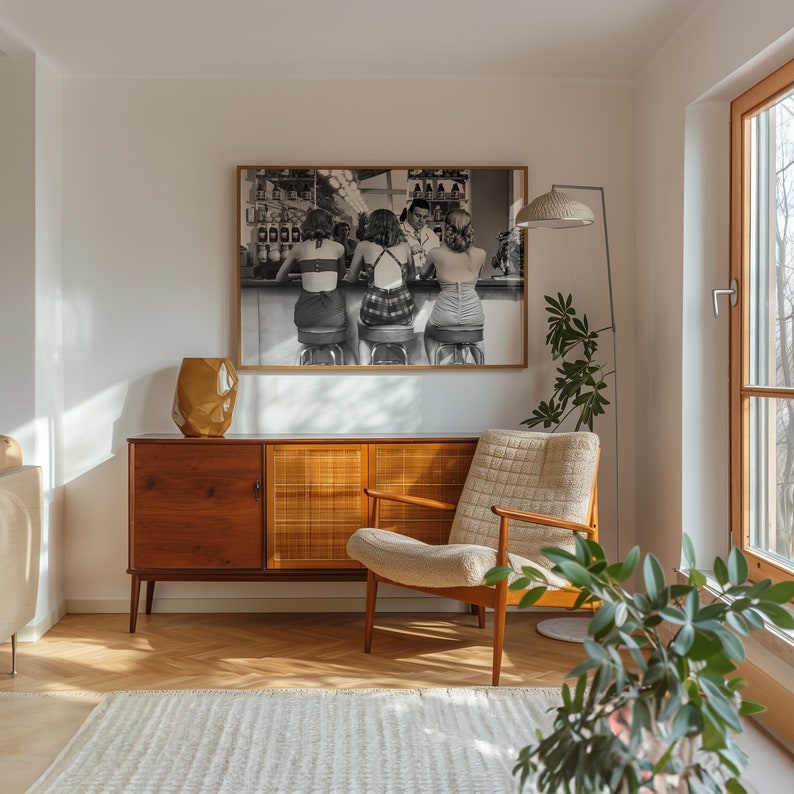 Op de afbeelding: Een woonkamer in mid-century modern stijl met een houten kast, een witte fauteuil en een zwart-witfoto van drie vrouwen die aan een bar zitten. De foto is ingelijst en hangt aan de muur. Er staat een plant in een pot in de hoek van de kamer.