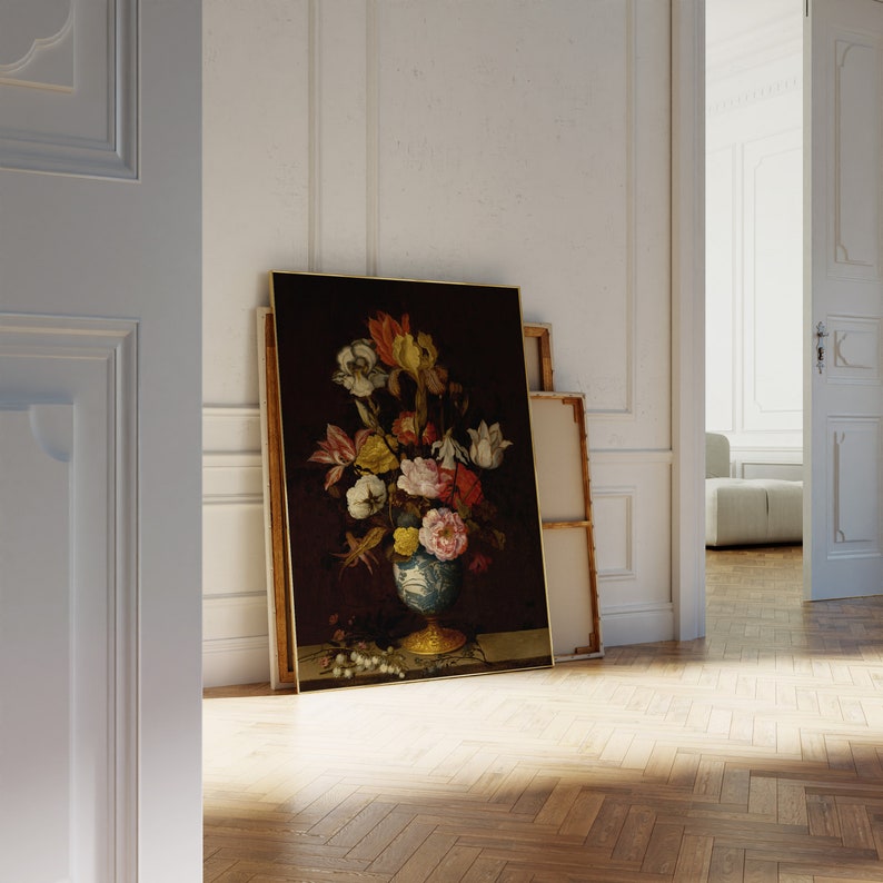 May include: A framed floral still life painting featuring a vase overflowing with colorful flowers. The vase is blue and white with a gold base. The painting leans against a white wall in a room with a wooden floor.