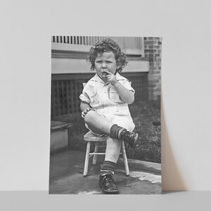 Peut inclure: Un jeune enfant aux cheveux boucl&eacute;s est assis sur une petite chaise en bois, portant une chemise blanche boutonn&eacute;e et des chaussures noires. Il tient un petit objet dans sa main et le regarde.