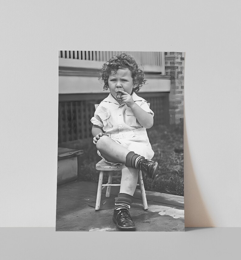 Peut inclure: Un jeune enfant aux cheveux boucl&eacute;s est assis sur une petite chaise en bois, portant une chemise blanche boutonn&eacute;e et des chaussures noires. Il tient un petit objet dans sa main et le regarde.
