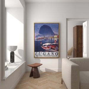 May include: A framed vintage travel poster for Lugano, Switzerland, depicting a lake scene with boats and a mountain backdrop. The poster is in a wooden frame and reads "LUGANO Suisse / Switzerland". The colour scheme includes blues, reds, and browns.