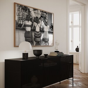 Op de afbeelding: Een zwart-witfoto van vier mensen die aan een bar zitten. De foto is ingelijst in een houten lijst en hangt aan een witte muur boven een zwarte kast met een lamp en decoratieve voorwerpen.