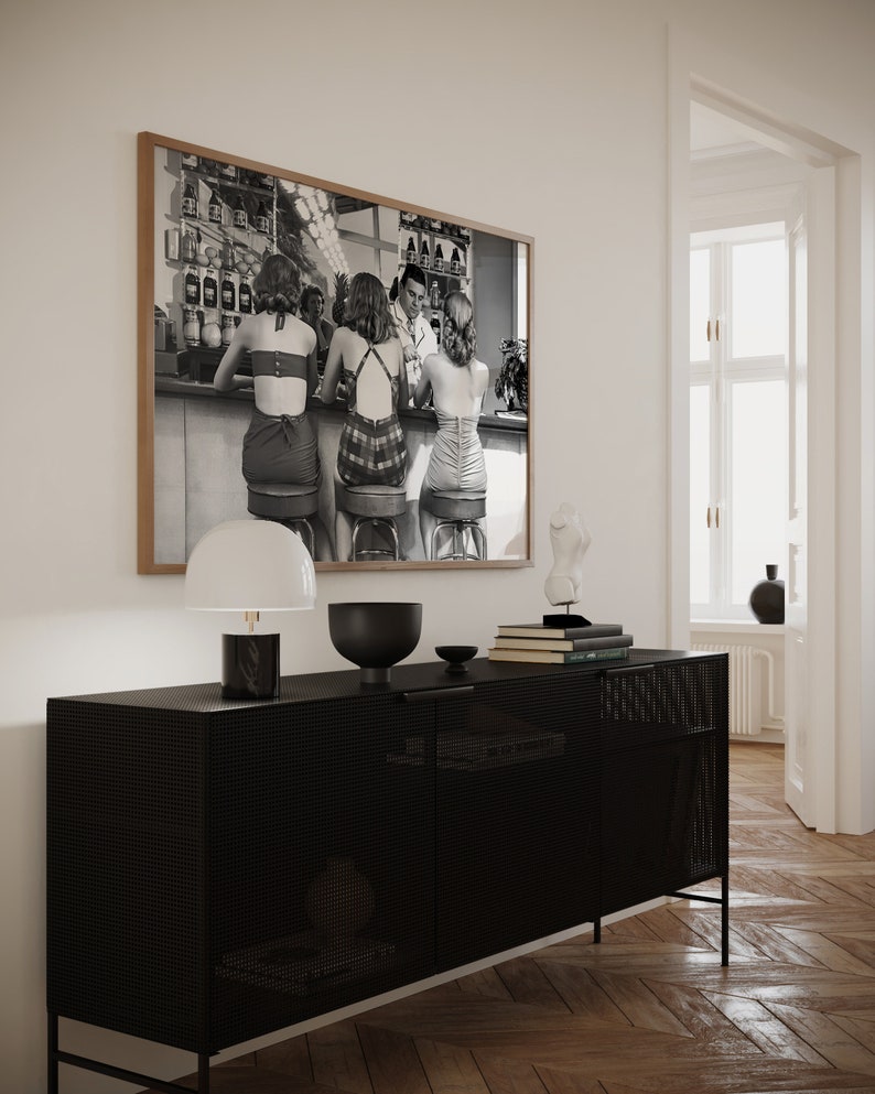 Op de afbeelding: Een zwart-witfoto van vier mensen die aan een bar zitten. De foto is ingelijst in een houten lijst en hangt aan een witte muur boven een zwarte kast met een lamp en decoratieve voorwerpen.