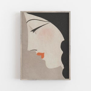 May include: Art Deco style print featuring a woman's profile in shades of gray, black, and peach. The woman's lips are a vibrant orange, and the artwork is framed in a light wood frame. A stylish piece of wall art.